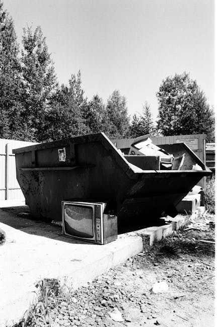 The image depicts a large, black industrial skip situated on a concrete paved area, likely part of a waste collection or disposal site. The skip is filled with various types of rubbish, including what