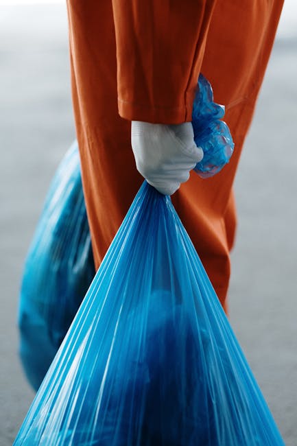 Close-up of a person wearing an orange protective suit and a white glove, holding a large blue plastic rubbish bag by the gathered top with their hand. The bag appears to be filled with waste material